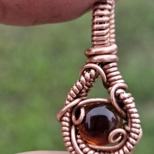 May include: A wire-wrapped pendant featuring a polished, round, amber-colored gemstone encased in a teardrop-shaped frame made of coiled copper wire. The metalwork consists of intricate, tightly wound copper strands that secure the central stone.