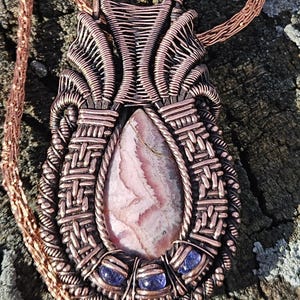 May include: A copper wire wrapped pendant with a pink and white stone and four small purple stones. The pendant is hanging from a copper chain.