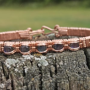 May include: A copper-colored wire-wrapped bracelet with five oval, dark purple gemstones. The bracelet is resting on a weathered, gray wooden surface, with a blurred green background.