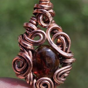May include: A pendant featuring a polished, oval-shaped dark amber gemstone encased in a decorative frame of coiled copper wire. The wire is wrapped in intricate loops around the stone, extending upward to form a secure bail for hanging.