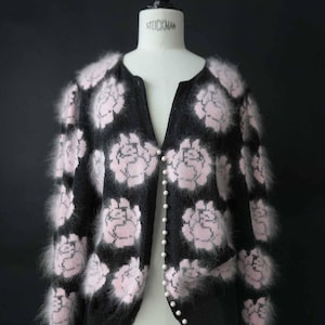 Könnte beinhalten: Schwarze Strickjacke mit rosa Blumenmuster. Der V-Ausschnitt-Pullover hat Perlenknöpfe und flauschige Verzierungen. Das Kleidungsstück wird auf einer Schneiderpuppe präsentiert.
