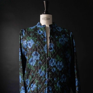 May include: A quilted coat with a floral pattern of blue roses and green leaves on a black background. The coat has a mandarin collar and is buttoned down the front with matching blue buttons. The coat is displayed on a mannequin.