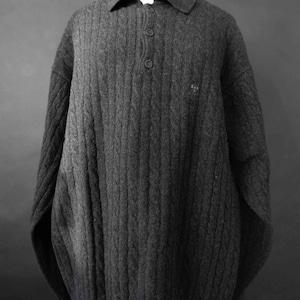 Peut inclure: Un pull en maille torsadée gris anthracite avec un col et trois boutons. Le pull présente un petit écusson brodé sur la poitrine. Le vêtement est présenté sur un mannequin sur fond gris foncé.