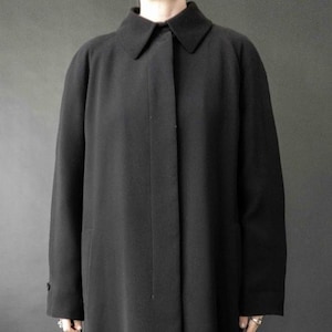 Könnte beinhalten: Ein langer, schwarzer Mantel mit klassischem Kragen und geknöpften Manschetten. Der Mantel ist einfarbig und scheint aus einem glatten Stoff zu bestehen. Der Mantel ist knielang und für kühleres Wetter geeignet.