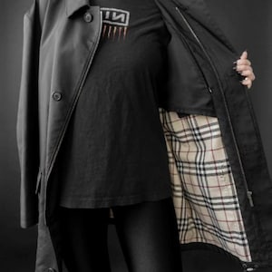 May include: Black trench coat with a button closure and a zipper. The coat's interior lining features a classic plaid pattern. The model is wearing a black t-shirt with a graphic design.