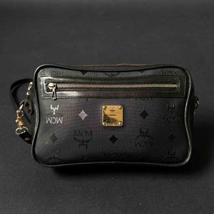 May include: Black crossbody bag with a repeating MCM logo pattern. The bag features a gold-toned zipper and hardware, and a small gold-colored rectangular emblem on the front. The bag has a long, adjustable strap.