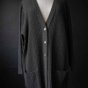 May include: A charcoal grey, ribbed-knit cardigan with a V-neck and long sleeves. The sweater has a button-up front, two front pockets, and is displayed on a mannequin. The cardigan is a versatile piece of clothing.