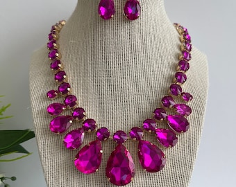 May include: A pink crystal necklace and earrings set with gold accents. The necklace features a teardrop design with multiple rows of smaller crystals.
