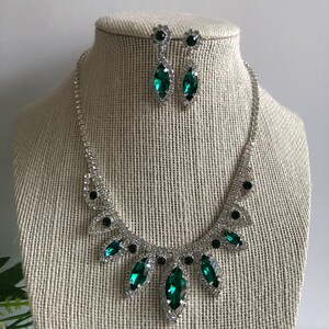 May include: A silver necklace and earrings set with emerald green and clear rhinestones. The necklace features a cascading design with five emerald green stones.