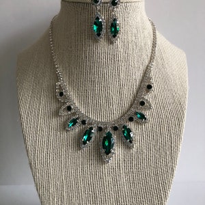 May include: A silver necklace and earrings set with emerald green rhinestones. The necklace features a teardrop design with a large emerald green rhinestone in the centre.