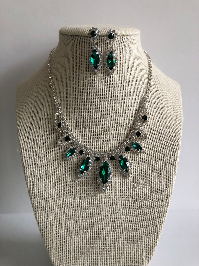 May include: A silver necklace and earrings set with emerald green rhinestones. The necklace features a teardrop design with a large emerald green rhinestone in the centre.