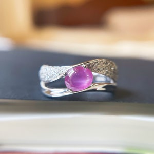 Op de afbeelding: Een zilveren ring met een roze edelsteen gezet in een gebogen band. De band is versierd met kleine diamanten.