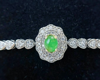 Op de afbeelding: Een zilveren armband met een hartvormig ontwerp en een grote groene edelsteen omringd door kleinere heldere edelstenen.