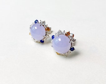 Puede incluir: Un par de pendientes de botón con piedras preciosas ovaladas de color lavanda. Cada pendiente está rodeado por un halo de pequeñas piedras preciosas blancas, azules y naranjas brillantes engastadas en metal plateado. Sobre fondo blanco.