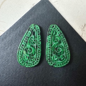 May include: Two green, teardrop-shaped pendants with intricate, swirling patterns and a beaded border. The pendants are displayed on a black and white surface, showcasing their detailed design and vibrant color.