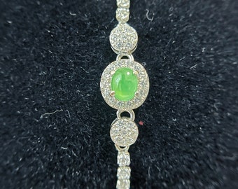 Op de afbeelding: Een zilveren armband met een groene edelsteen omringd door kleine heldere stenen. De armband heeft een delicaat kettingontwerp.
