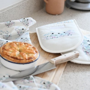 May include: A pair of white oven gloves with a watercolour design of sheep in a field. The gloves have a blue and white striped border and the text "The Country Rose" is printed on one glove.