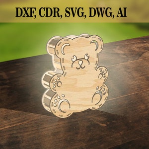 Puede incluir: Un recorte de madera de un oso de peluche marrón con un diseño cosido. El oso tiene una cara simple con dos ojos, una nariz y una boca.