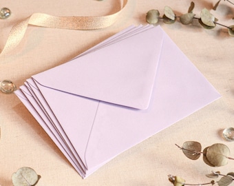 May include: A stack of pale lavender envelopes, arranged on a neutral-toned surface. The envelopes are fanned out, showcasing their triangular flap design. Decorative elements include a gold ribbon, clear glass beads, and sprigs of greenery, suggesting stationery or invitation supplies.
