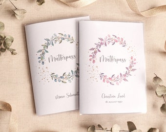 May include: Two white booklets with floral wreath designs. One features a blue and green wreath with the word "Mutterpass" and the name "Marie Schmidt". The other has a pink wreath, the same word, and the name "Christina Lund".