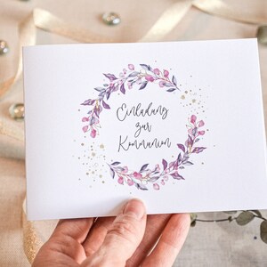 May include: A white card with a purple and pink floral wreath with gold glitter accents. The text "Einladung zur Kommunion" is written in cursive inside the wreath.