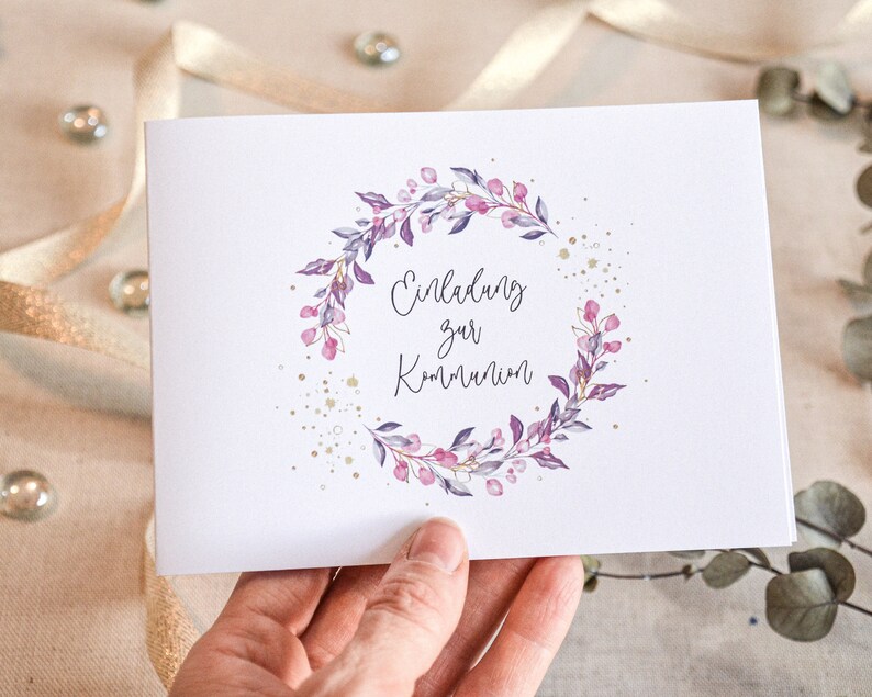 May include: A white card with a purple and pink floral wreath with gold glitter accents. The text "Einladung zur Kommunion" is written in cursive inside the wreath.