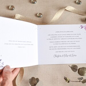 May include: A white card with purple floral accents and gold glitter. The text reads "Liebe Familie & Freunde, Wir m&ouml;chten euch ganz herzlich zur Taufe unserer Leone am 15.06.2022 um 11 Uhr in der St. Stephan Kirche in Hamburg einladen. Anschliessend freuen wir uns mit euch in der Pulverm&uuml;hle zu feiern. Bitte gebt uns bis zum 01.03.2023 Bescheid, ob ihr den besonderen Tag mit uns feiert. Wir freuen uns auf euch Christine & Felix at Leone"