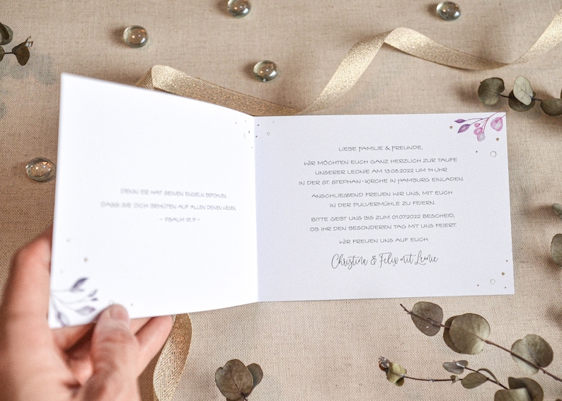 May include: A white card with purple floral accents and gold glitter. The text reads "Liebe Familie & Freunde, Wir m&ouml;chten euch ganz herzlich zur Taufe unserer Leone am 15.06.2022 um 11 Uhr in der St. Stephan Kirche in Hamburg einladen. Anschliessend freuen wir uns mit euch in der Pulverm&uuml;hle zu feiern. Bitte gebt uns bis zum 01.03.2023 Bescheid, ob ihr den besonderen Tag mit uns feiert. Wir freuen uns auf euch Christine & Felix at Leone"