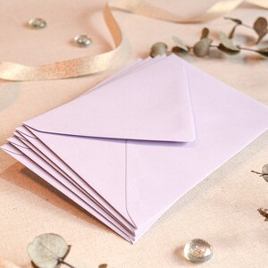 May include: A stack of light purple envelopes on a beige fabric background. The envelopes are folded and ready to be used for sending letters or cards.