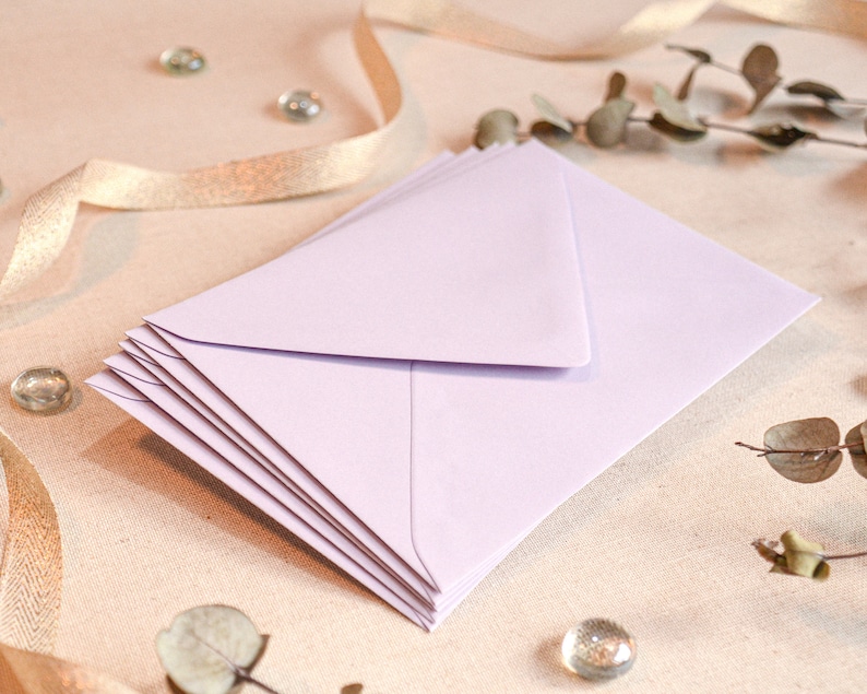 May include: A stack of light purple envelopes on a beige fabric background. The envelopes are folded and ready to be used for sending letters or cards.