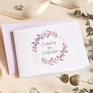 May include: A white card with a purple floral wreath and gold glitter. The text on the card reads "Einladung zur Konfirmation".