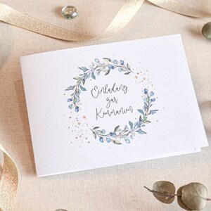 May include: A white card with a blue and green floral wreath and gold glitter. The text on the card reads "Einladung zur Kommunion".