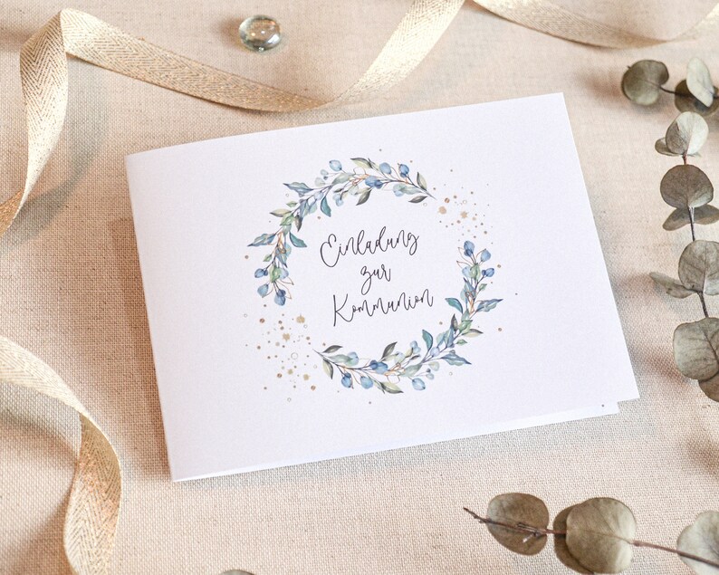 May include: A white card with a blue and green floral wreath and gold glitter. The text on the card reads "Einladung zur Kommunion".