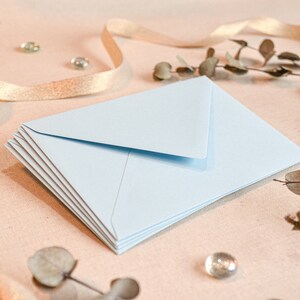 May include: A stack of light blue envelopes on a beige surface with gold ribbon and dried foliage.