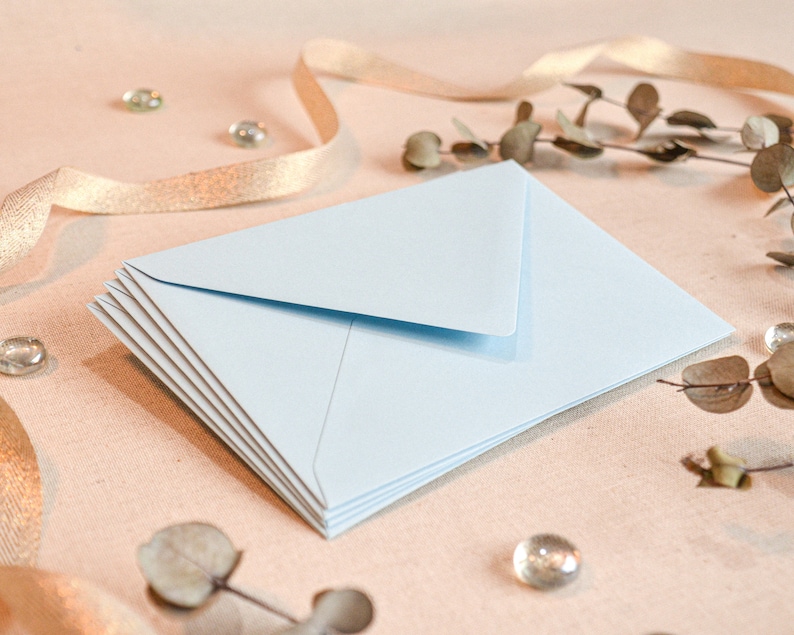 May include: A stack of light blue envelopes on a beige surface with gold ribbon and dried foliage.