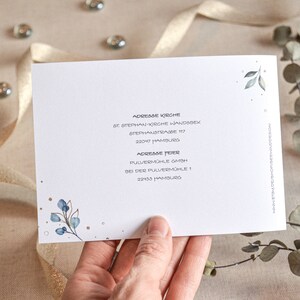 May include: A white card with blue and green watercolor leaves and gold glitter. The card has the text "Adresse Kirche" and "Adresse Feier" with addresses for St. Stephan-Kirche Wandsbek and Pulverm&uuml;hle GmbH.