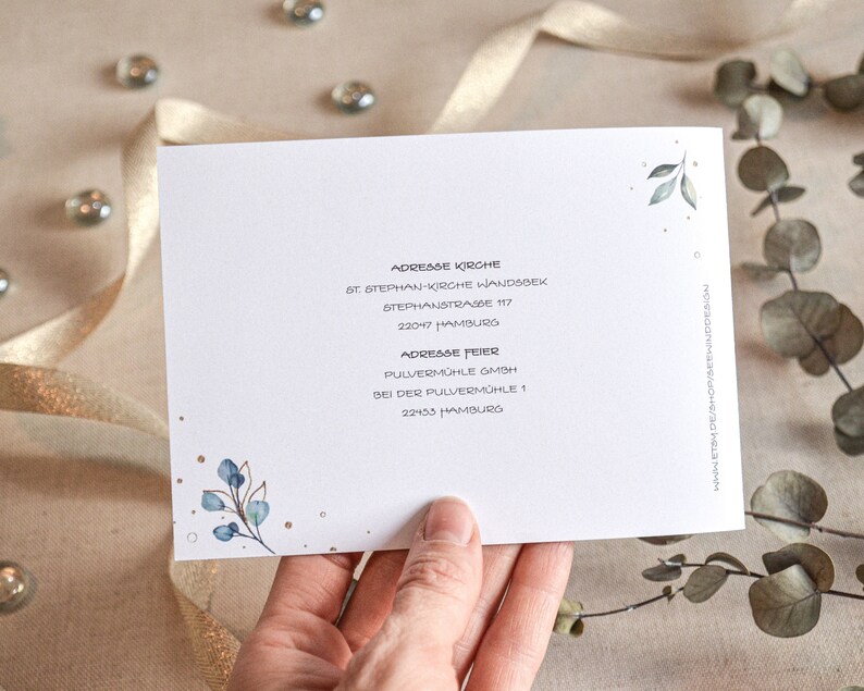May include: A white card with blue and green watercolor leaves and gold glitter. The card has the text "Adresse Kirche" and "Adresse Feier" with addresses for St. Stephan-Kirche Wandsbek and Pulverm&uuml;hle GmbH.