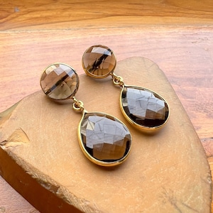 May include: A pair of gold-toned earrings with faceted, smoky quartz gemstones. The earrings feature a round stud and a teardrop-shaped dangle, both set in gold. The gemstones have a warm, brown hue and are displayed on a wooden surface.