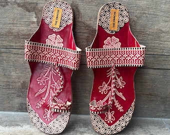 May include: A pair of red leather sandals with white floral designs and black circular patterns. The sandals have a strap across the top of the foot and a toe loop. The sandals are labeled "THE ENACHE" on the straps.