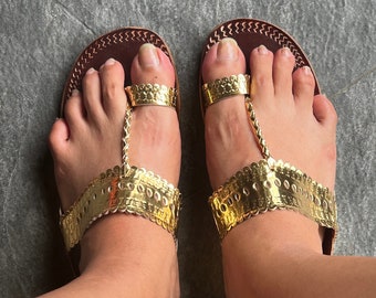 May include: A pair of gold and brown leather sandals with a toe ring and decorative stitching. The sandals are worn on a person's feet.