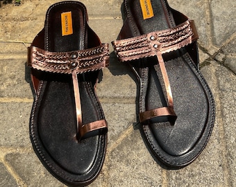 May include: A pair of brown leather sandals with a gold metallic trim and a toe ring. The sandals have a brand label that says "The Enachie".