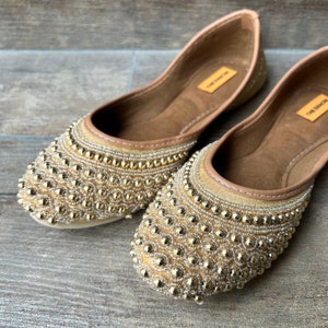 May include: A pair of beige flat shoes with gold and silver bead embellishments. The shoes have a pointed toe and a flat sole. The brand name "The  Shoe" is visible on the insole.