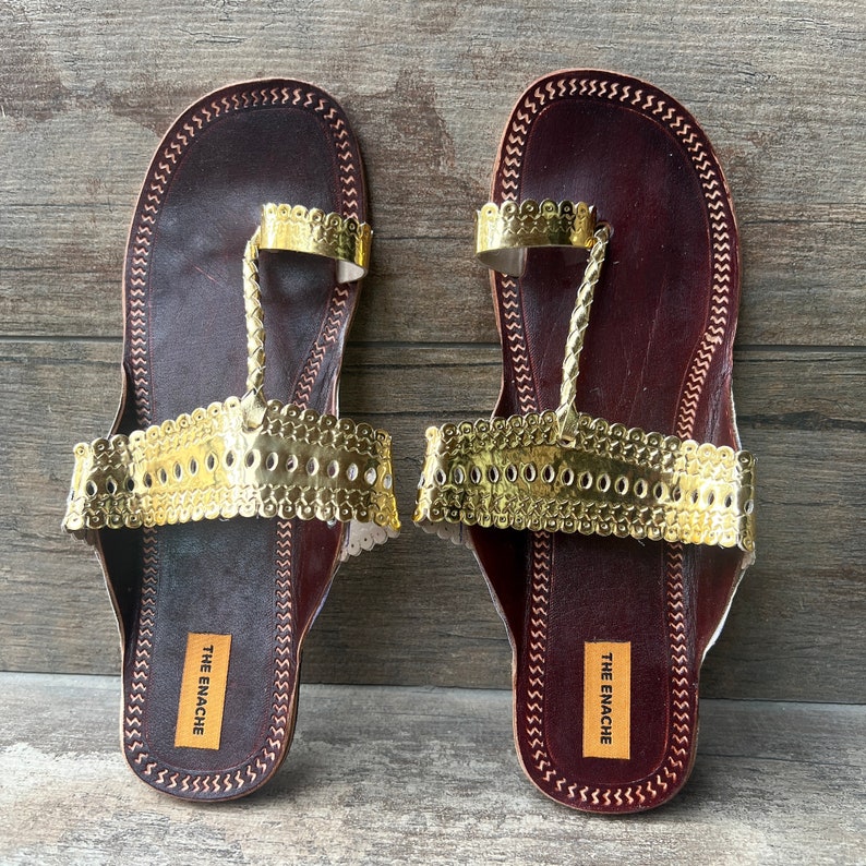 May include: A pair of brown leather sandals with gold-colored embellishments. The sandals have a toe strap and an ankle strap. The brand name "THE ENACHE" is printed on the sole of each sandal.