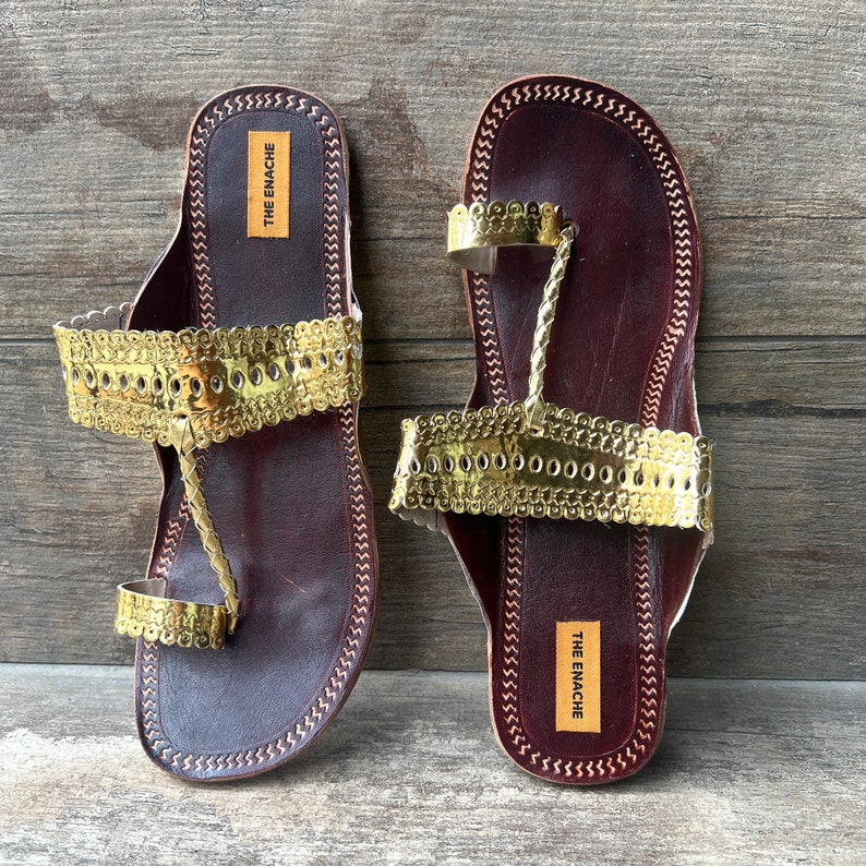 May include: A pair of brown leather sandals with gold embellishments. The sandals have a strap that goes around the ankle and a toe ring. The sandals have the text "THE ENACHE" on the sole.