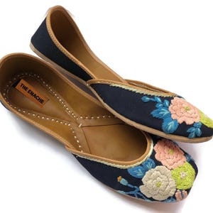 Puede incluir: Un par de bailarinas negras con bordados florales en rosa, azul y verde. Los zapatos tienen suelas y ribetes de cuero marr&oacute;n. La marca "THE ENACHE" es visible en la plantilla.