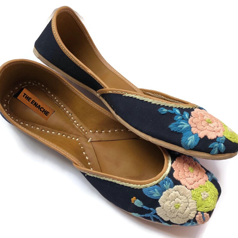 Puede incluir: Un par de bailarinas negras con bordados florales en rosa, azul y verde. Los zapatos tienen suelas y ribetes de cuero marr&oacute;n. La marca "THE ENACHE" es visible en la plantilla.