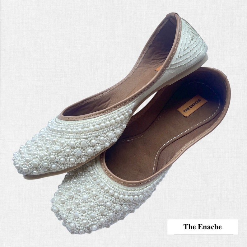 May include: A pair of white flat shoes with pearl embellishments. The shoes have a brown leather insole and are made by The Enachie.