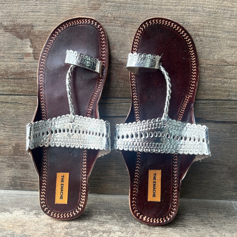 May include: A pair of brown leather sandals with silver embellishments. The sandals have a toe loop and an ankle strap. The sandals are labeled "The Enachie".