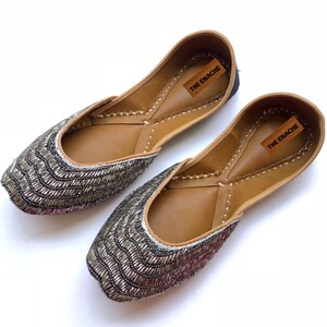 Peut inclure: Une paire de ballerines orn&eacute;es d'un motif de perles argent&eacute;es sur le bout et les c&ocirc;t&eacute;s. Les chaussures ont un int&eacute;rieur en cuir marron et une &eacute;tiquette qui dit "THE ENACHE". Les ballerines sont sur fond blanc.