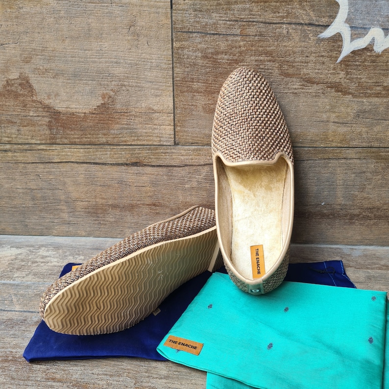 Op de afbeelding: Een paar bruine geweven instappers met een textuurzool. De schoenen liggen op een blauwe en groene stof met de tekst "THE ENACHE" erop gedrukt.